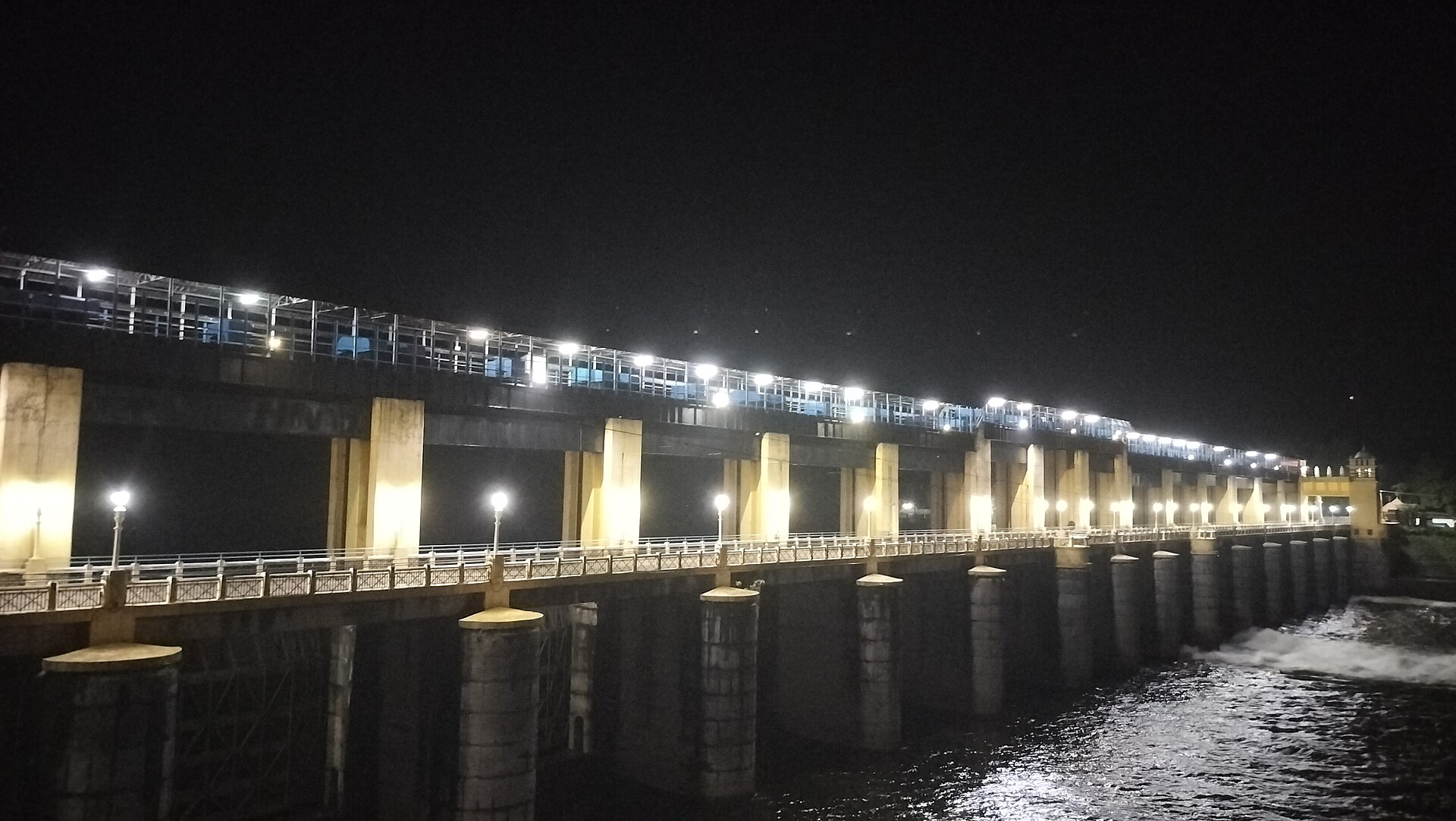 एर्णाकुलम के भूतत्तानकेट्टु बांध से रात में पानी छोड़ा जा रहा है, जून 2026 की तस्वीर, फाटकों के नीचे सफ़ेद धार साफ़ दिखती है। जिन नदियों पर ऐसे बांध हैं, वहां छोड़े गए पानी का फ़ैसला कोई इंसान लेता है।