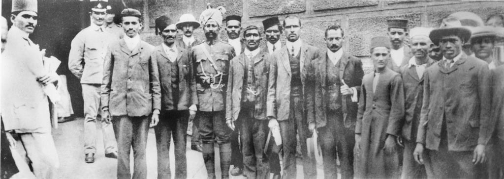 दक्षिण अफ़्रीका में जेल के बाहर खड़े गांधी और उनके साथी, भीड़ में वर्दी पहने दो लोग भी हैं, यह तस्वीर 1908 की है; एम्पायर थिएटर के प्रस्ताव में जेल जाने की जो बात तय हुई थी, उसके क़रीब दो साल बाद का यह दृश्य है