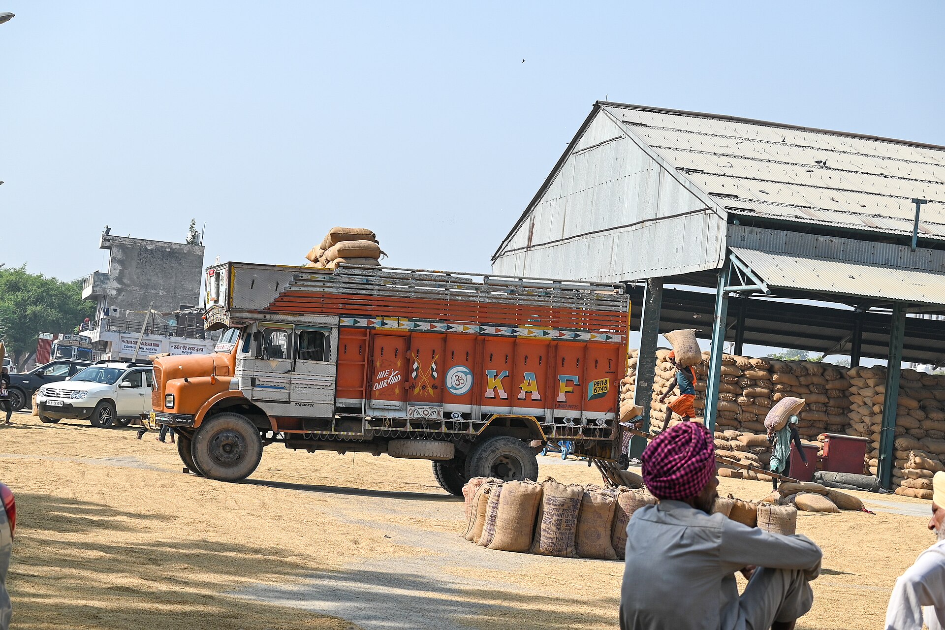 पंजाब की एक अनाज मंडी में अनाज की बोरियाँ ट्रक पर लादी जा रही हैं और फ़र्श पर बिखरा अनाज पड़ा है, अक्टूबर 2023 की तस्वीर। थोक टोकरी का खाद्य हिस्सा ऐसी ही मंडियों में बने भाव से बनता है